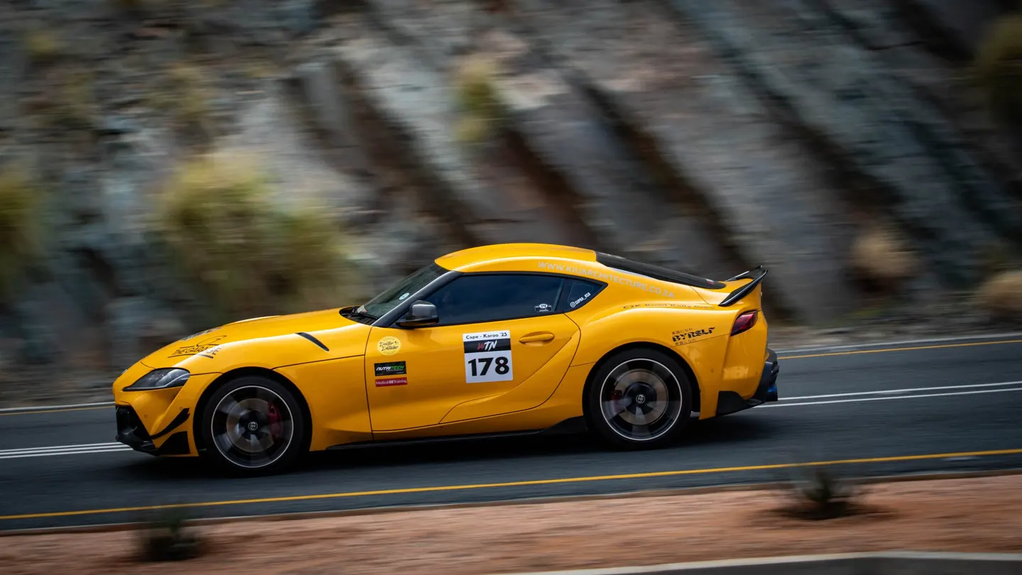 A yellow Toyota Supra at speed past a rock face on Cape to Karoo