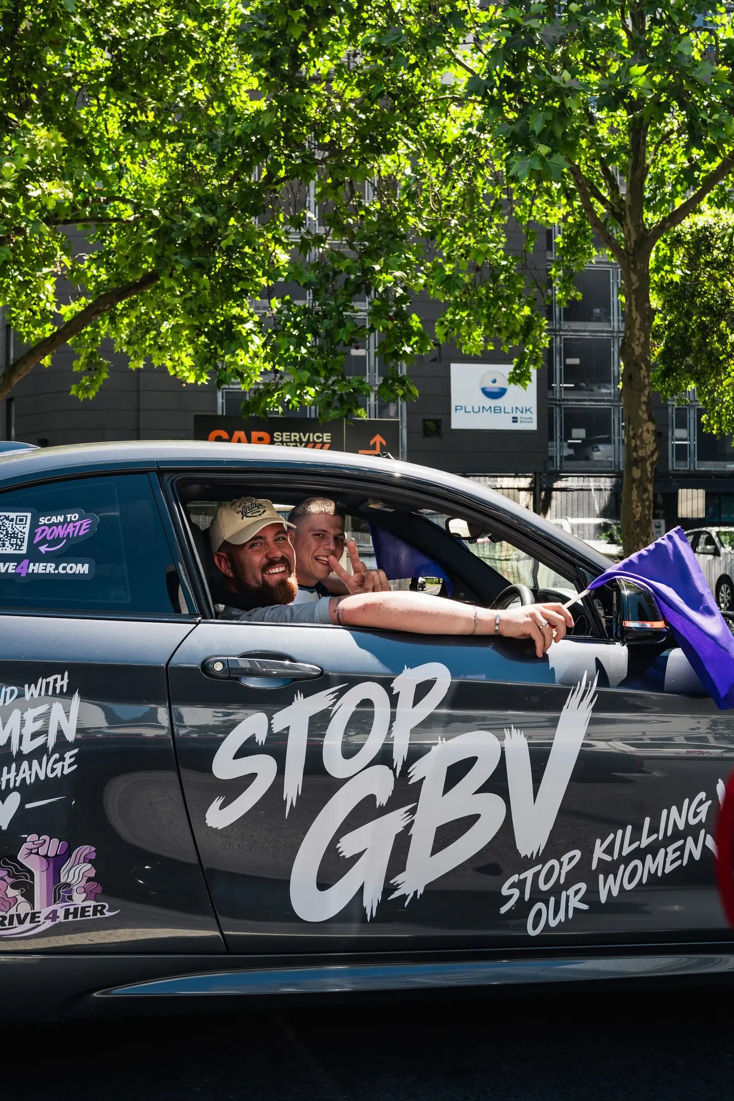 A driver and passenger waving a purple flag from a Drive4Her car